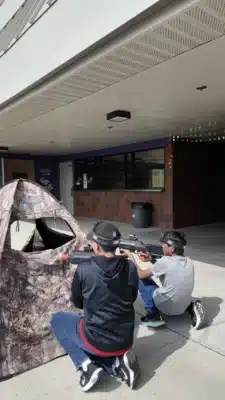 2 kids kneeling behind a bunker holding laser tag guns on concrete with a high school outside entrance in the background