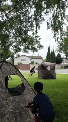 kid hiding behind a bunker playing laser tag and a kid running in the background with another bunker in a residential neighborhood