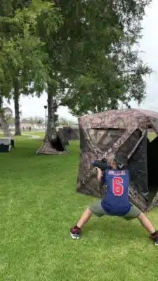 teen aiming laser tag gun with laser tag bunkers in the background. green grass public park and bunkers underneath tree with lots of leaves