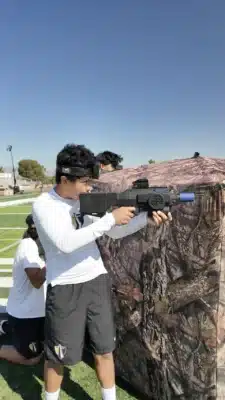 teen aiming a laser tag gun wearing a white shirt and grey shorts taking cover behind a camo bunker playing laser tag on a football field with a track meet in the background with blue sky